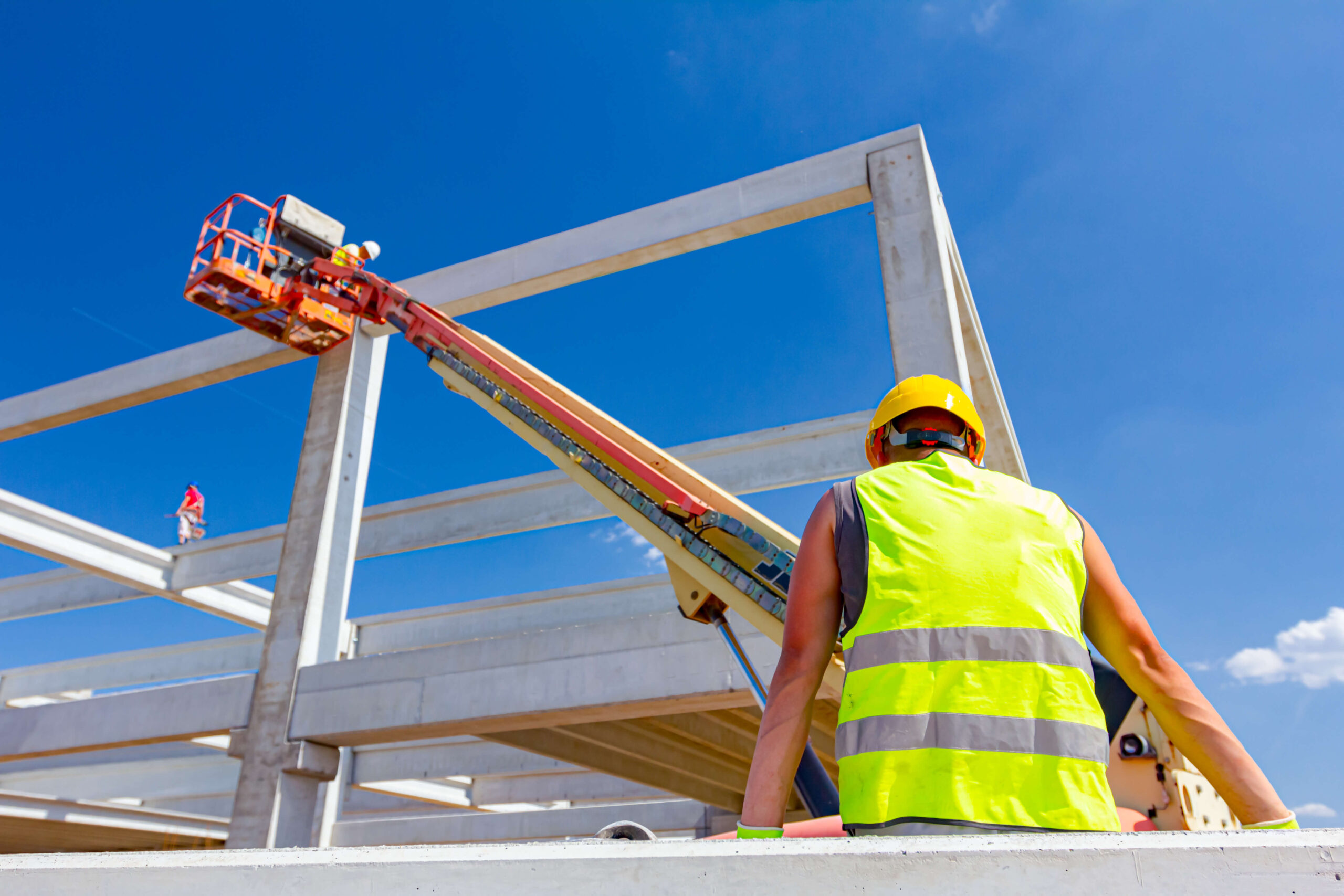 Arbeidere bruker lift for å montere betongelementer på byggeplass under klar blå himmel, iført verneutstyr og refleksvester.